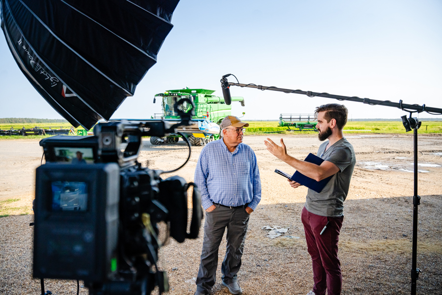 A film crew is interviewing a farmer in front of a combine harvester on a sunny day.